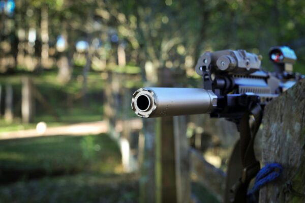 A detailed view of a rifle with a silencer in a wooded area, focus on the firearm barrel.